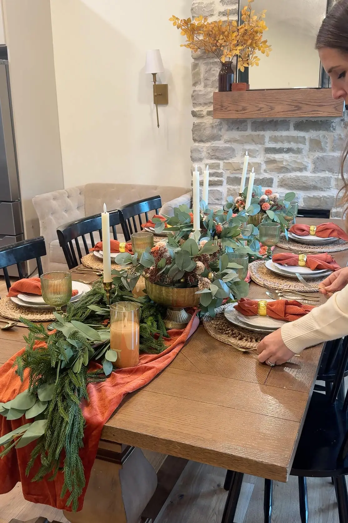 Holiday dining table set with plates, woven placemats, orange napkins, and a centerpiece of greenery, candles, and floral arrangements.